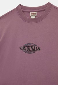 Paarse katoenen T-shirt met een ronde halslijn en een zwart "ORIGINALS GUESS" graphic print op de borst, met een wereldbol ontwerp.