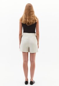 Pantaloni corti beige chiaro su misura con una texture liscia, orlo risvoltato, tasche laterali e vita a media altezza, abbinati a una canotta nera attillata.