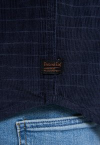 Marineblauwe stof shirt met horizontale ribbeling, voorzien van een bruine label met de tekst "Petrol Ind" op de zoom, gecombineerd met blauwe denim.