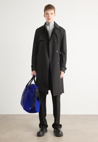 Black double-breasted trench coat, grey high-neck top, black trousers, and black shoes. Holding a blue waterproof duffel bag with black straps.