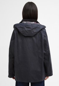 Casaco encerado azul-marinho com capuz, apresentando um interior com forro de xadrez e um corte descontraído. Exterior texturizado com rugas visíveis.