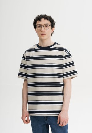 Jonge man met krullend haar en bril, die een beige en zwarte horizontaal gestreepte t-shirt en blauwe jeans draagt, staand tegen een eenvoudige witte achtergrond.