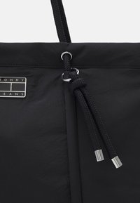 Svart tygpåse med flätat dragsnöre knutet i en knut och silverfärgade metallsnören, med en rektangulär logotavla för varumärket.