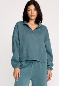 Teal pullover sweatshirt, cropped design, halvdels-lynlås foran, ribbede manchetter, børstet tekstur, med et lille logo på brystet.