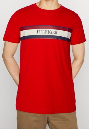 Mann trägt ein rotes kurzärmliges T-Shirt mit einem horizontalen weißen und marineblauen Streifen und dem Schriftzug "HILFIGER", kombiniert mit beigen Hosen.