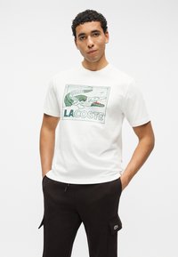 Νεαρός με σγουρά μαλλιά, που φοράει λευκό t-shirt Lacoste με πράσινο κροκόδειλο και μαύρο παντελόνι με πλαϊνές τσέπες.