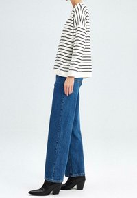 Person, der blaue weite Jeans, schwarze Absatzstiefel und einen lockeren weißen Pullover mit schwarzen horizontalen Streifen trägt und seitlich steht.