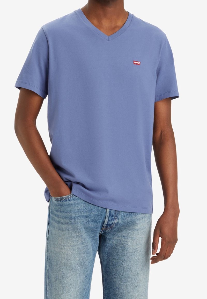 Homme portant un T-shirt Levi’s bleu clair à col en V et un jean denim bleu clair, une main dans la poche, sur fond blanc.