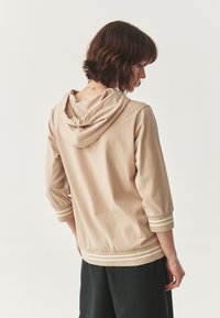 Sudadera beige con capucha, mangas de tres cuartos, puños a rayas y dobladillo acanalado. Textura suave, ajuste casual.