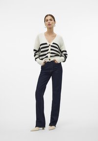 Striped knit cardigan in cream and black with large buttons, paired with dark blue straight-leg jeans and beige pointed-toe heels.