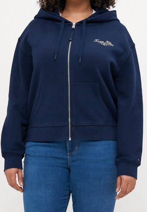 Person wearing navy blue zip-up hoodie with front pockets and Tommy Hilfiger logo, paired with blue jeans, against a plain background.