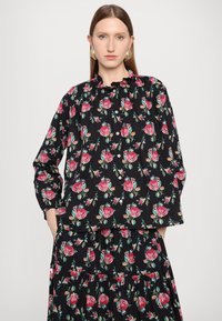 Svart blomstret bluse med rosa og teal blomster, volangkrage, knappelukking og lange ermer. Kombinert med et matchende mønstret skjørt.