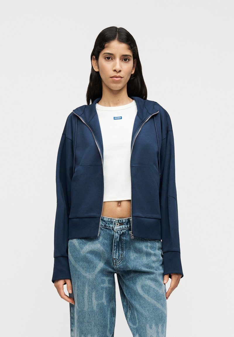 Μπλε σκούρο φούτερ με φερμουάρ από μαλακό ύφασμα με άνετη εφαρμογή, που διαθέτει κουκούλα, δύο μπροστινές τσέπες και ριμπ μανσέτες, συνδυασμένο με τζιν ανοιχτού μπλε χρώματος.