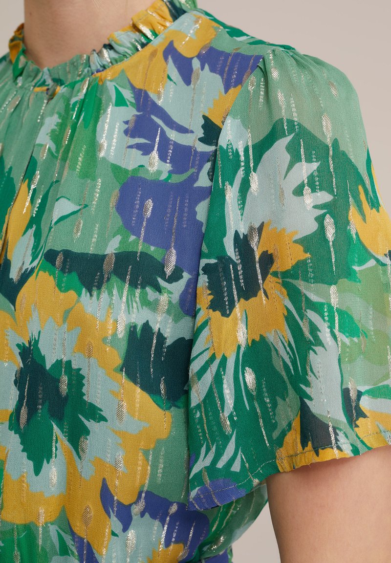 Kurzärmlige Bluse mit grünem Blumenmuster und gelben sowie blauen Akzenten, aus leichtem Stoff gefertigt und mit metallischen Streifen strukturiert.