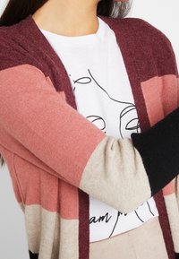 Stickad cardigan med vinröda, rosa, krämfärgade och svarta ränder; mjuk textur; öppen framdesign; lager på en vit t-shirt med grafiskt tryck.