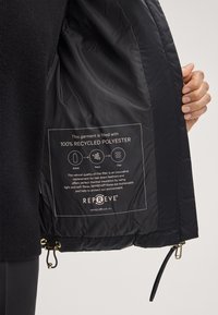 Schwarze isolierte Jacke mit Innenfutter aus 100 % recyceltem Polyester. Verfügt über ein Etikett, das das Material erläutert, sowie einen Reißverschlussdetails.