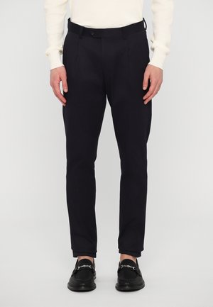 Man draagt zwarte slim-fit broek, zwarte loafers met zilveren kettingen en een crèmekleurige trui, staand tegen een effen achtergrond.