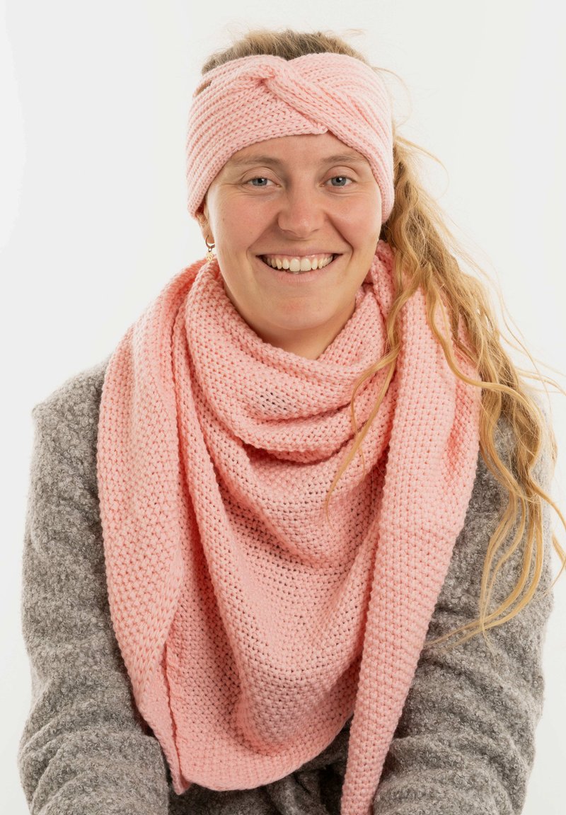 Femme souriante avec de longs cheveux blonds, portant un bandeau en tricot rose et une écharpe, ainsi qu'un pull en maille grise sur fond uni.
