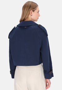 Kvinna som bär en marinblå kort jacka med knäppta axelband och cremefärgade högmidjade byxor, sedd bakifrån mot en vit bakgrund.