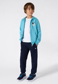 Felpa blu chiaro con zip e grafica di un astronauta sul petto, abbinata a pantaloni della tuta blu navy con un piccolo logo. Sneakers nere e blu.