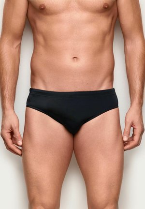 Atletyczny tors mężczyzny w czarnych kąpielówkach, stojący na jednolitym jasnym tle z rękami swobodnie opuszczonymi wzdłuż ciała.