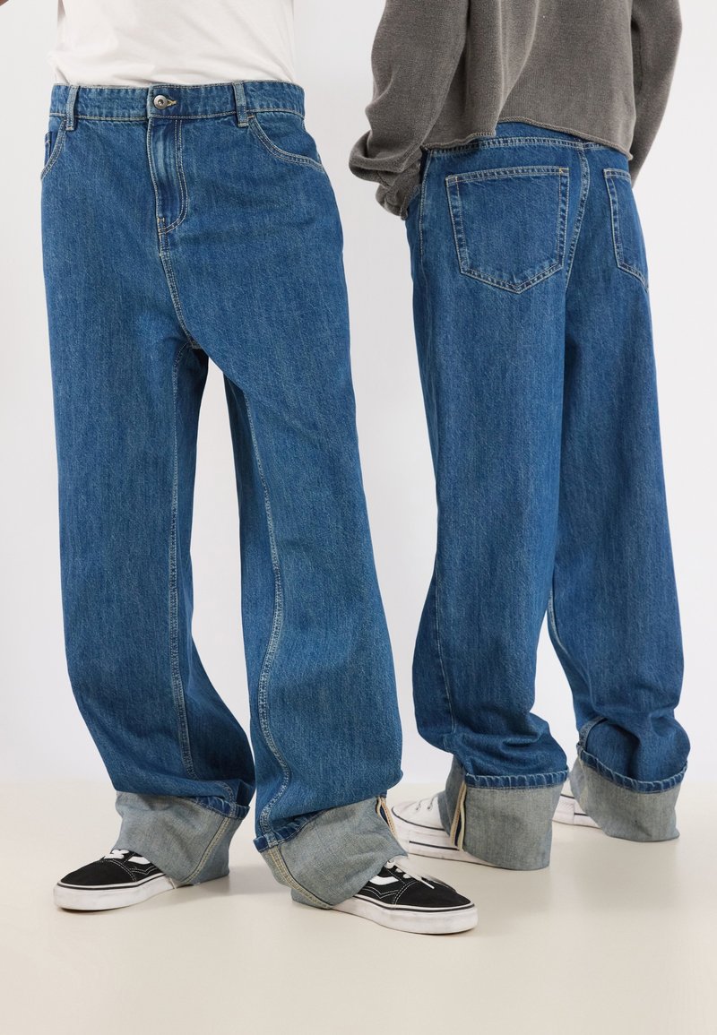 Deux personnes portant des jeans bleus surdimensionnés avec de grands revers, associés à des baskets décontractées, debout devant un fond uni.