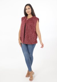 Weste aus Kunstfell ohne Ärmel in Burgunder, mit weicher Textur und offenem Vorderteil; kombiniert mit blauen Skinny-Jeans und klaren High-Heels.