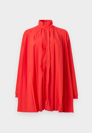 Giacca rossa brillante a maniche lunghe con colletto alto, cerniera frontale e design ampio plissettato, mostrata su uno sfondo bianco semplice.