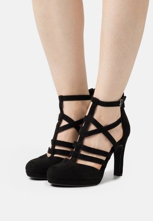 Zwarte suède hoge hakken met een gesloten neus, uitgevoerd met gekruiste bandjes en een enkelriempje met gesp. De hakhoogte is ongeveer 10 centimeter.