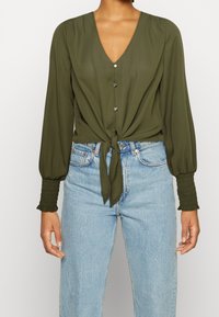 Blusa verde oliva con scollo a V, bottoni frontali e nodo in vita. Presenta maniche lunghe e puffate con polsini arricciati.