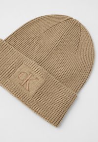 Beidzis adīts beanie krēmkrāsā ar rievotu faktūru, salocītu apakšmalu un izšuvumu brūnā krāsā ar logo plāksteri, kurā ir burts "K" priekšpusē.