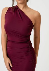 Bordeaux sukienka na jedno ramię wykonana z elastycznego materiału, z drapowaniem po bokach i gładką fakturą. Brak widocznych elementów metalowych lub wzorów.