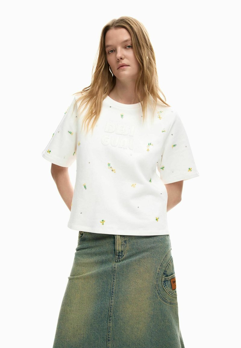 Mujer joven con pelo largo y rubio, lleva una camisa blanca de manga corta con pequeños estampados florales y una falda vaquera desgastada con un parche en el bolsillo.