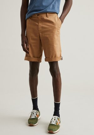 Shorts en coton beige avec un ourlet retroussé, des poches avant et une fermeture à bouton, associés à des baskets vertes et des chaussettes foncées.