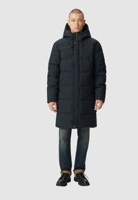 Giacca a vento blu scuro con cappuccio, dotata di sezioni trapuntate, zip frontale e bottoni a pressione. Indossata sopra jeans in denim e scarpe nere.