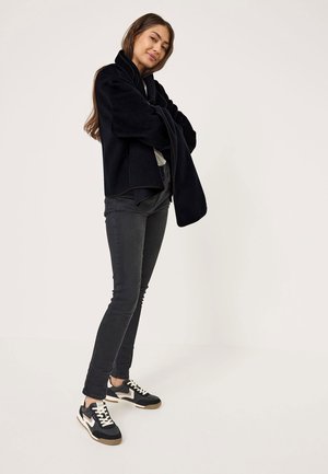 Black fleece jacket with shawl collar, paired with fitted gray jeans and black sneakers featuring white accents and beige soles.