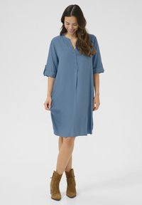Abito blu, fino alle ginocchia, realizzato in tessuto liscio, con colletto, chiusura a bottoni e maniche arrotolate. Abbinato a stivaletti alla caviglia color beige.