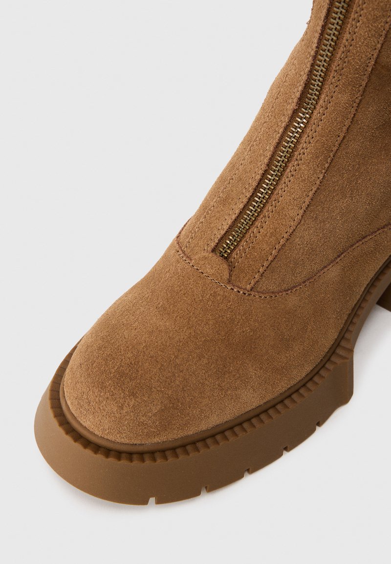 Bruine suède enkelboot met een ronde neus, zijrits, gestructureerde rubberen zool en stiksels. Heeft een gladde, matte afwerking.