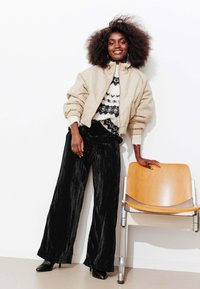 Veste bomber beige, pantalon large noir texturé, pull à motifs avec des accents blancs et noirs, et bottes à talons noires, sur un fond neutre.