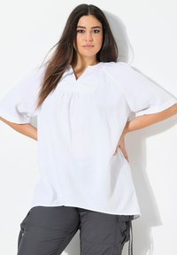 Top bianco e testurizzato con scollatura a V e maniche corte, caratterizzato da dettagli arricciati sotto il colletto. Abbinato a pantaloni cargo scuri.