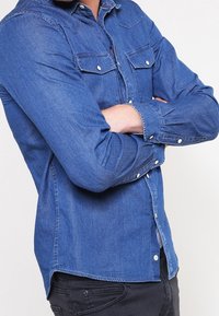 Tummansininen farkkupaita, pitkät hihat, napit edessä, kaksi rintataskua läpillä ja kontrastoivaa ompelua saumoissa.