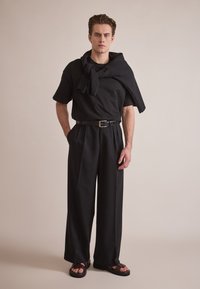 Svart t-skjorte med nakkesløyfe, høyt liv svart vide bukser og svarte sandaler. Glatt stoff med matt finish og minimalistisk design.