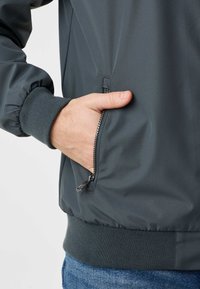 Χέρι στην πλαϊνή τσέπη ενός γκρι ανθρακί μπουφάν με φερμουάρ, με ριμπ μανσέτα και τελείωμα, φορεμένο πάνω από μπλε τζιν, σε ανοιχτό φόντο.