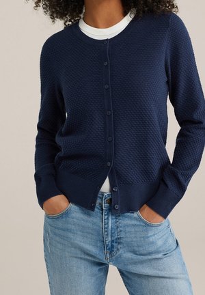Vrouw draagt een marineblauwe, gestructureerde, knoopsluitende cardigan over een wit shirt, met handen in de zakken van lichtblauwe spijkerbroek.