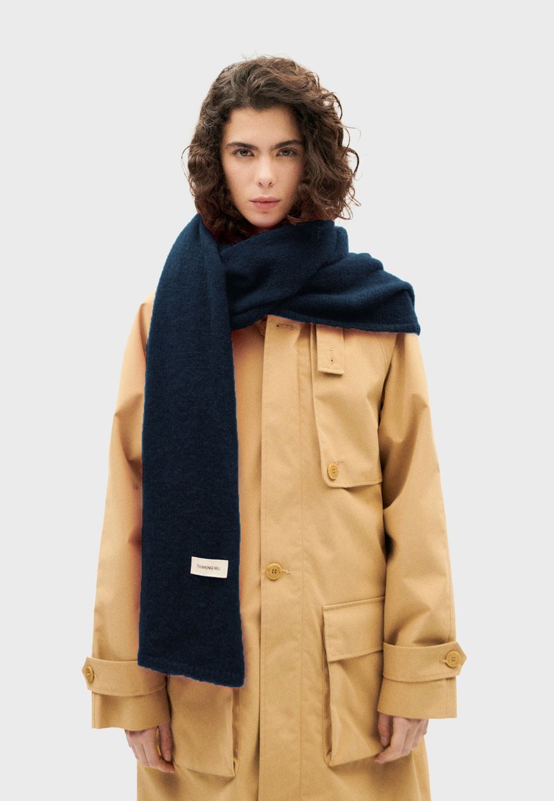 Marineblaues Wollschal, rechteckige Form, weiche Textur, locker um den Hals drapiert, mit einem Markenetikett an der Seite.