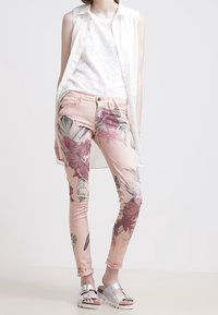 Roze skinny jeans met bloemenpatroon en groene accenten, gecombineerd met een witte textuur tanktop en een lange, lichte crèmevest. Zilveren sandalen.