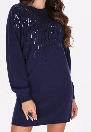 Femme portant une robe pull bleu marine à manches longues ornée de sequins noirs et argentés sur le haut de la poitrine et les manches, tenant une grosse bague sombre.