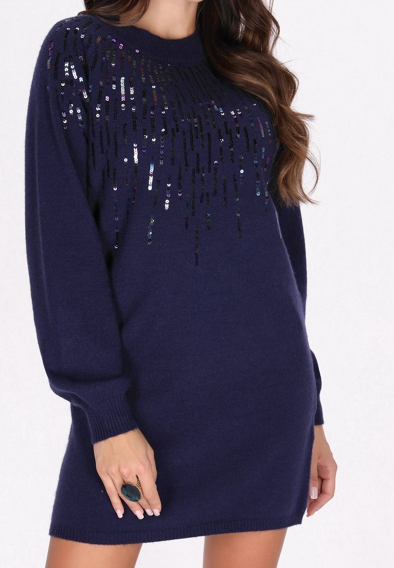 Femme portant une robe pull bleu marine à manches longues ornée de sequins noirs et argentés sur le haut de la poitrine et les manches, tenant une grosse bague sombre.