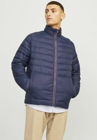 Marineblå puffjakke med quilted tekstur, høj krave og kontrastfarvet lynlås. Båret over en lyseblå stribet skjorte og beige bukser.