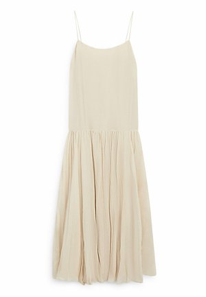 Femme avec les cheveux attachés en chignon, portant une robe sans manches beige clair, regardant vers le bas et sur le côté sur un fond gris clair uni.